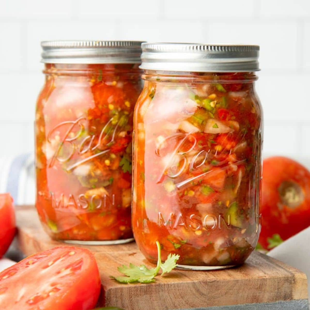 Two mason jars filled with salsa sit on a wooden cutting board with tomatoes around them