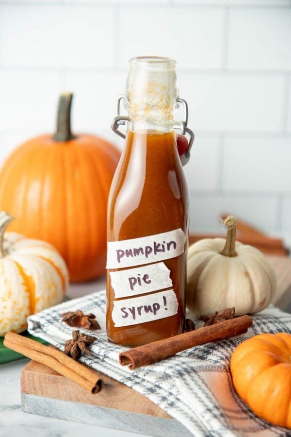 A flip-top glass bottle filled with pumpkin spice syrup sits among decorative pumpkins, cinnamon sticks, and whole star anise.