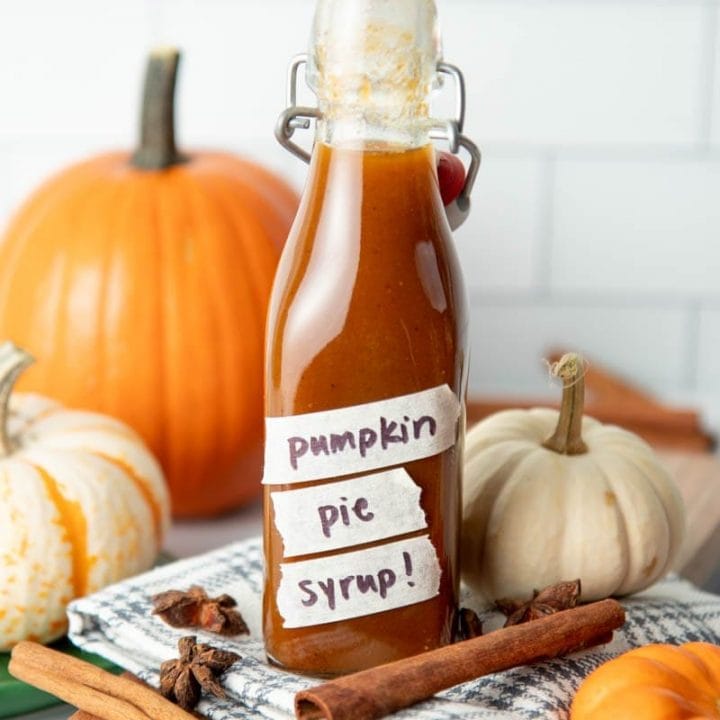 A flip-top glass bottle filled with pumpkin spice syrup sits among decorative pumpkins, cinnamon sticks, and whole star anise.