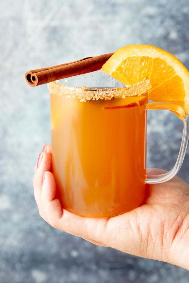 Close-up of hand cupping the bottom of a mug of hot apple cider garnished with a sugar rim, orange slice, and cinnamon stick.