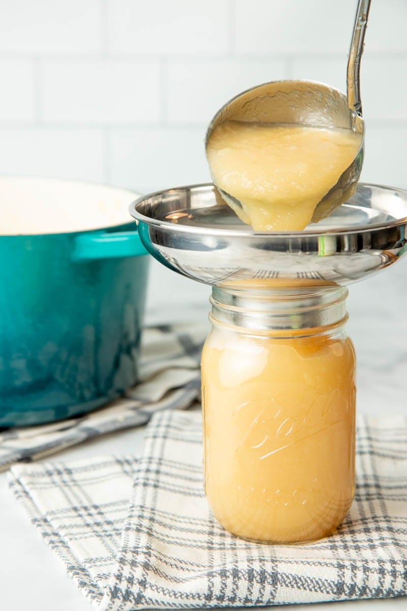 A ladle pours homemade applesauce through a canning funnel into a pint jar for canning.