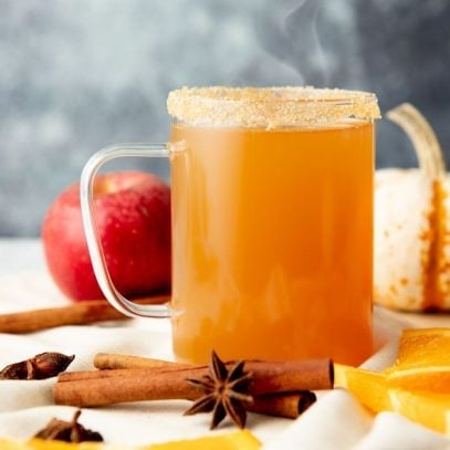 A mug of apple cider spiked with bourbon sits with steam rising from the top surrounded by orange slices, whole spices, an apple and a pumpkin.