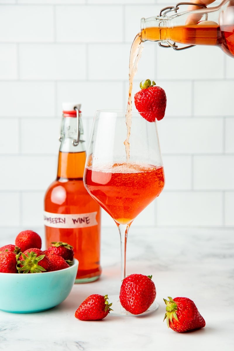 Pouring a glass of homemade strawberry wine with a fresh strawberry garnish on the glass.