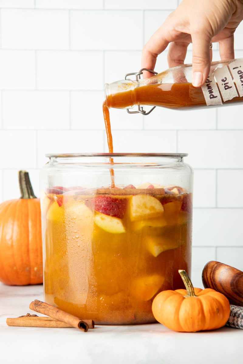 A hand pours pumpkin spice syrup into a large pitcher.