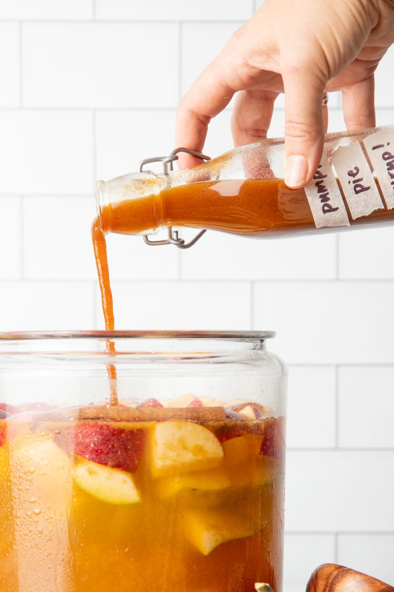 Close-up of a hand pouring pumpkin spice syrup into large glass pitcher of pumpkin pie sangria.