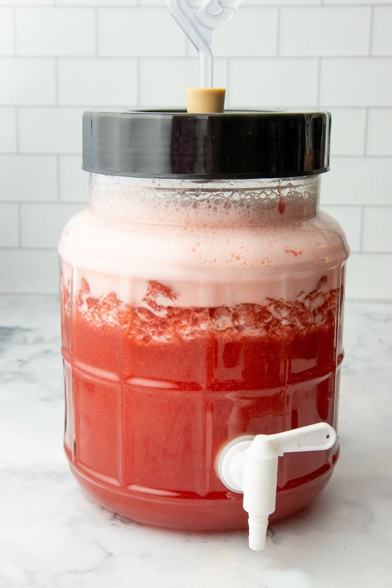 A glass fermenter with a spigot is filled with vigorously bubbling strawberry wine in the primary fermentation stage.