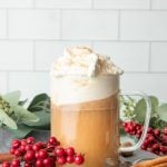 A hand sprinkles a pinch of nutmeg over the whipped cream topping a mug of hot buttered rum.