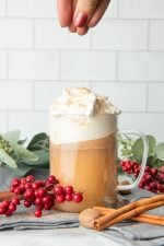 A hand sprinkles a pinch of nutmeg over the whipped cream topping a mug of hot buttered rum.
