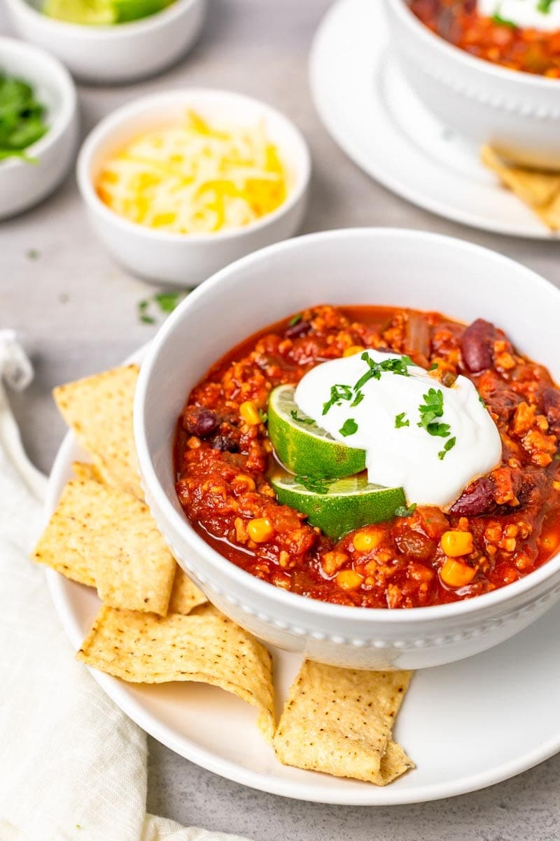 A full bowl of taco turkey chili garnished with lime, sour cream, and cilantro sits on a plate with tortilla chips.