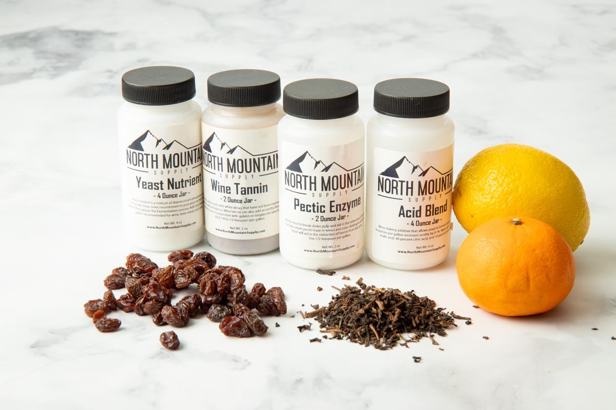 Bottles of wine additives from North Mountain Supply such as yeast nutrients, wine tannin, pectic enzyme, and acid blend along with fresh additives like raisins, citrus, and tea.