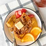 Overhead of a single serving of overnight french toast topped with butter and maple syrup on a plate surrounded by fresh strawberries and orange slices.