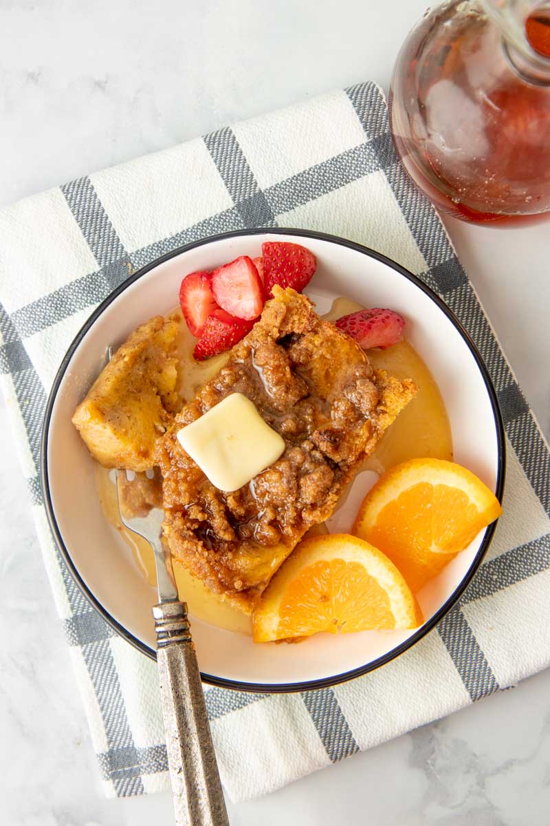 Overhead of a single serving of overnight french toast topped with butter and maple syrup on a plate surrounded by fresh strawberries and orange slices.