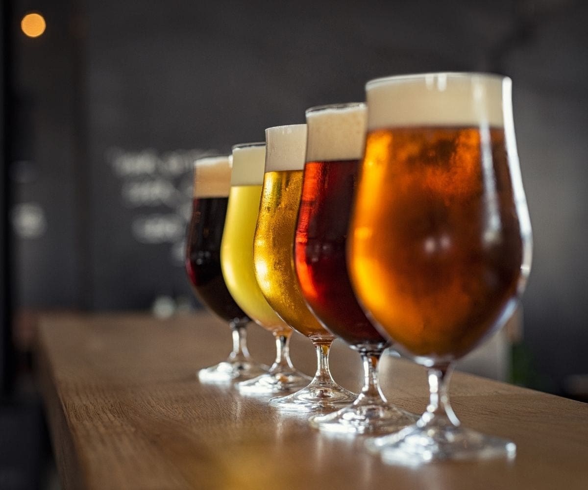 Five tulip beer glasses lined up along a bar and filled with beers in different hues.