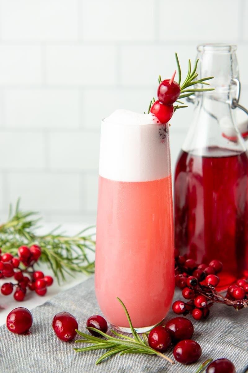 A mixed cranberry gin fizz cocktail stands beside an open flip-top bottle of cranberry simple syrup.