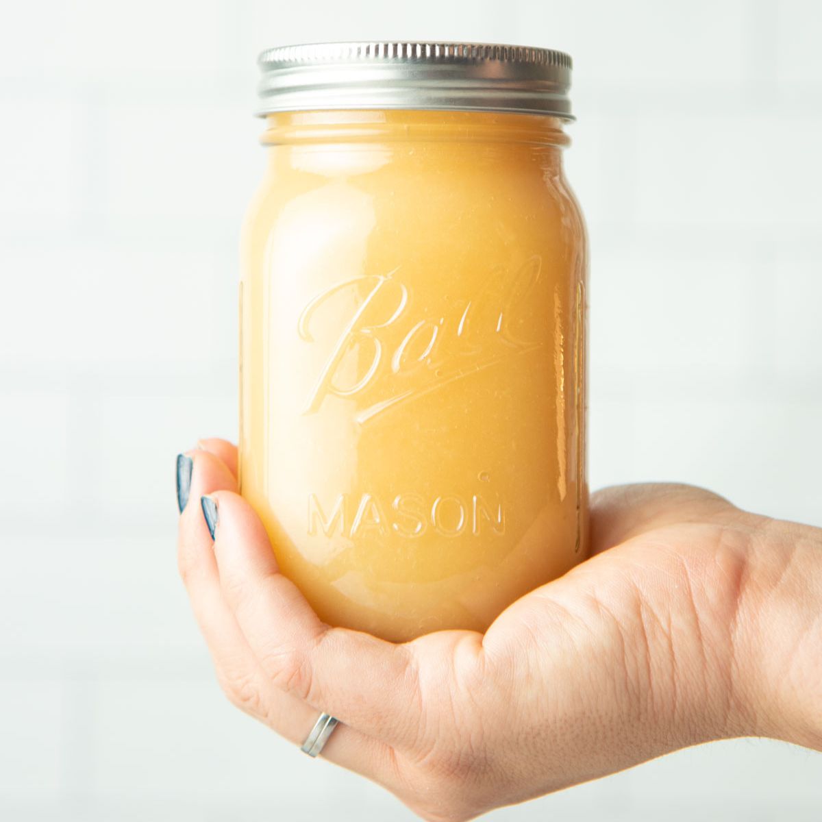 Hand holds a jar of finished canned applesauce in front of a white tile backsplash