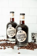 Two bottles of homemade kahlua on a kitchen counter surrounded by coffee beans.