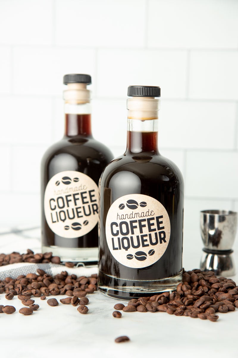 Two bottles of homemade kahlua on a kitchen counter surrounded by coffee beans. Liqueurs like this make great Christmas food gifts!