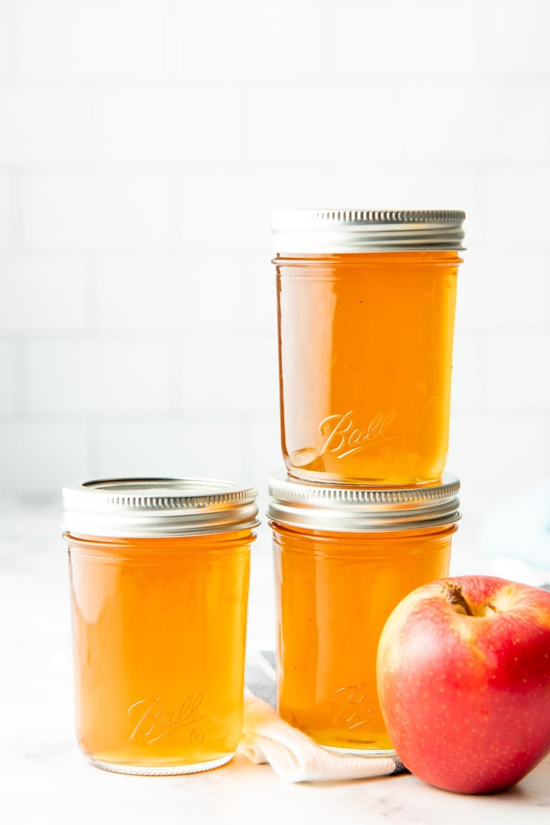 Three pint-sized jars of apple jelly stacked next to a fresh apple.