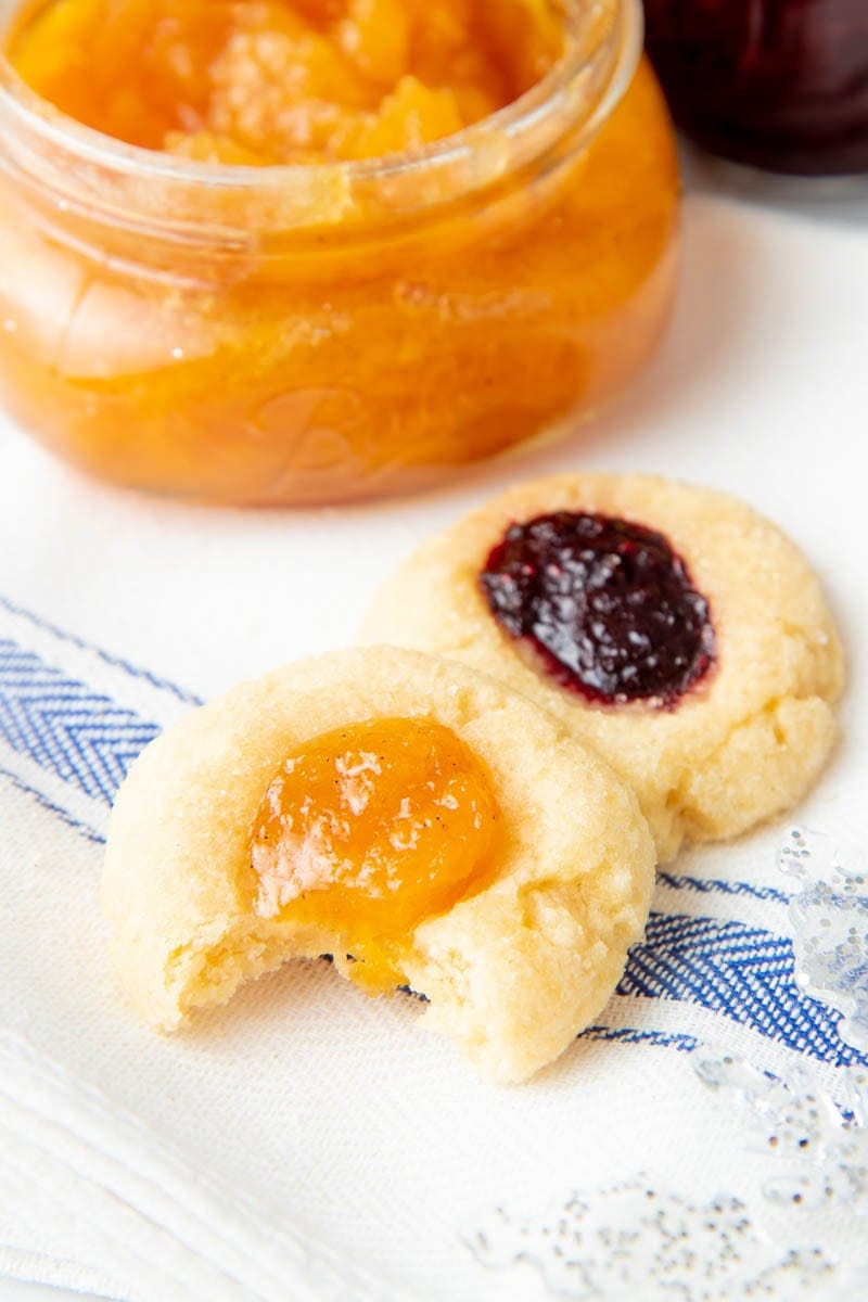 Close-up of a jam thumbprint cookie with a bite out of it sitting in front of another cookie and an open jar of jam.