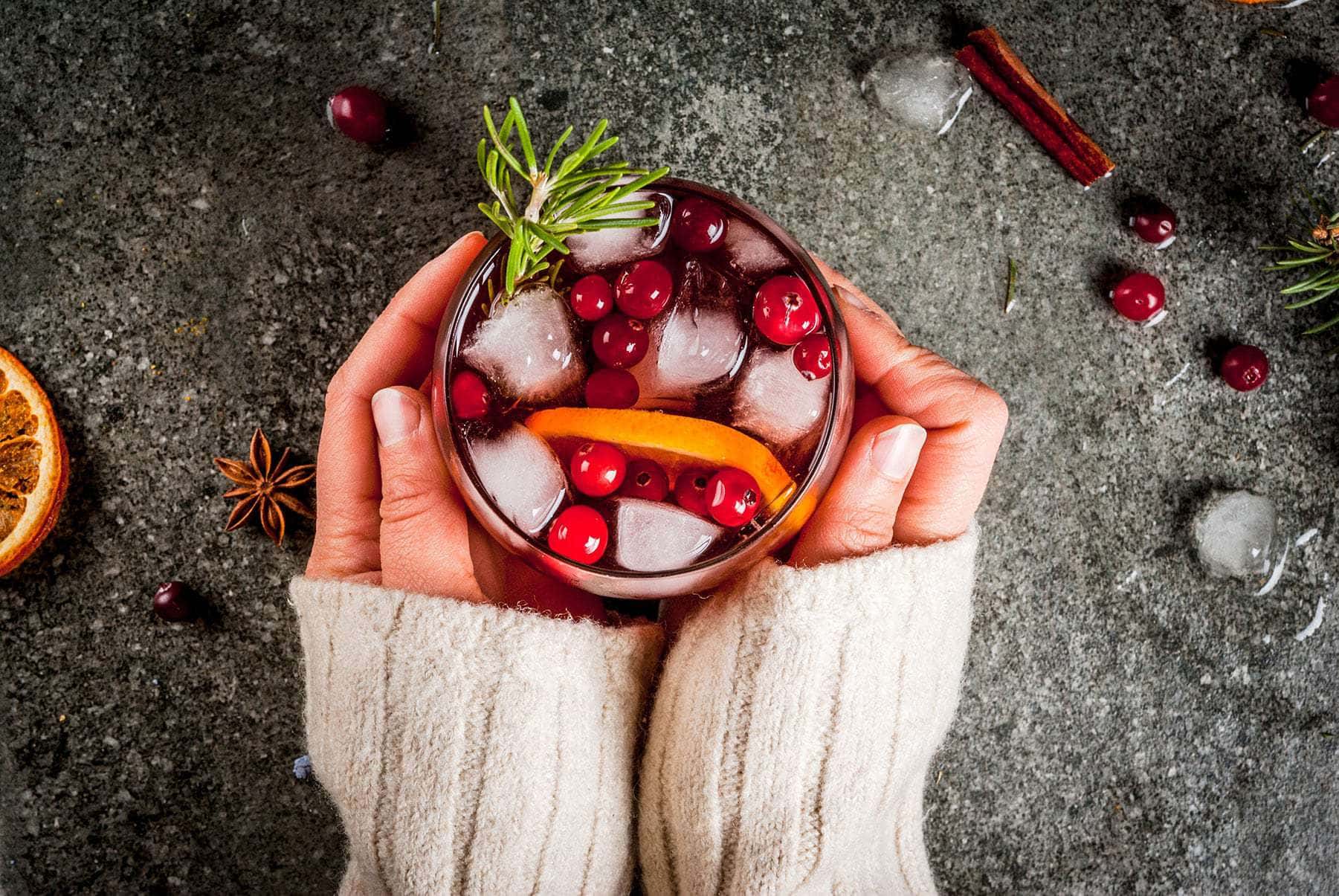 Top view of two hands holding a festive christmas cocktail garnished with cranberries, an orange slice, and a rosemary sprig.