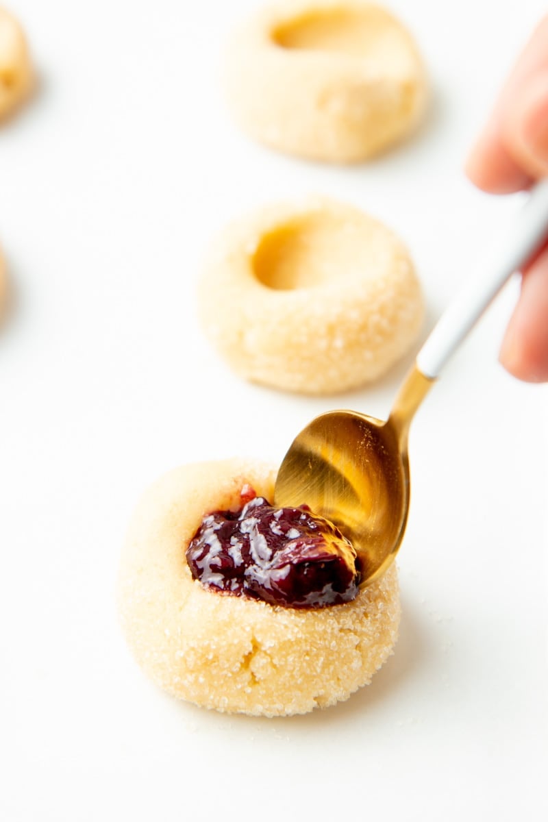 Close-up of spooning jam into the thumbprint on top of a cookie.