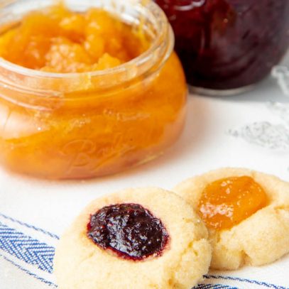 Close-up of two jam thumbprint cookies with open jars of homemade jam behind them.