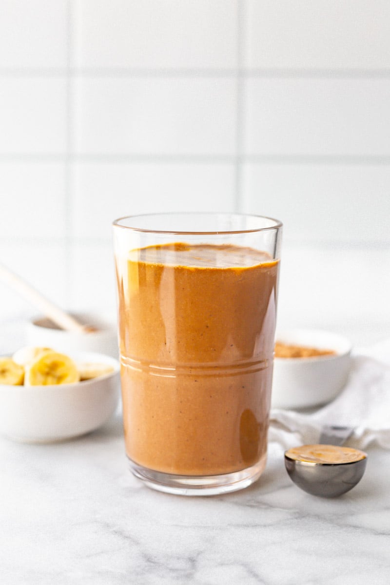 A chocolate peanut butter smoothie sits in front of small bowls filled with the ingredients to make the smoothie.