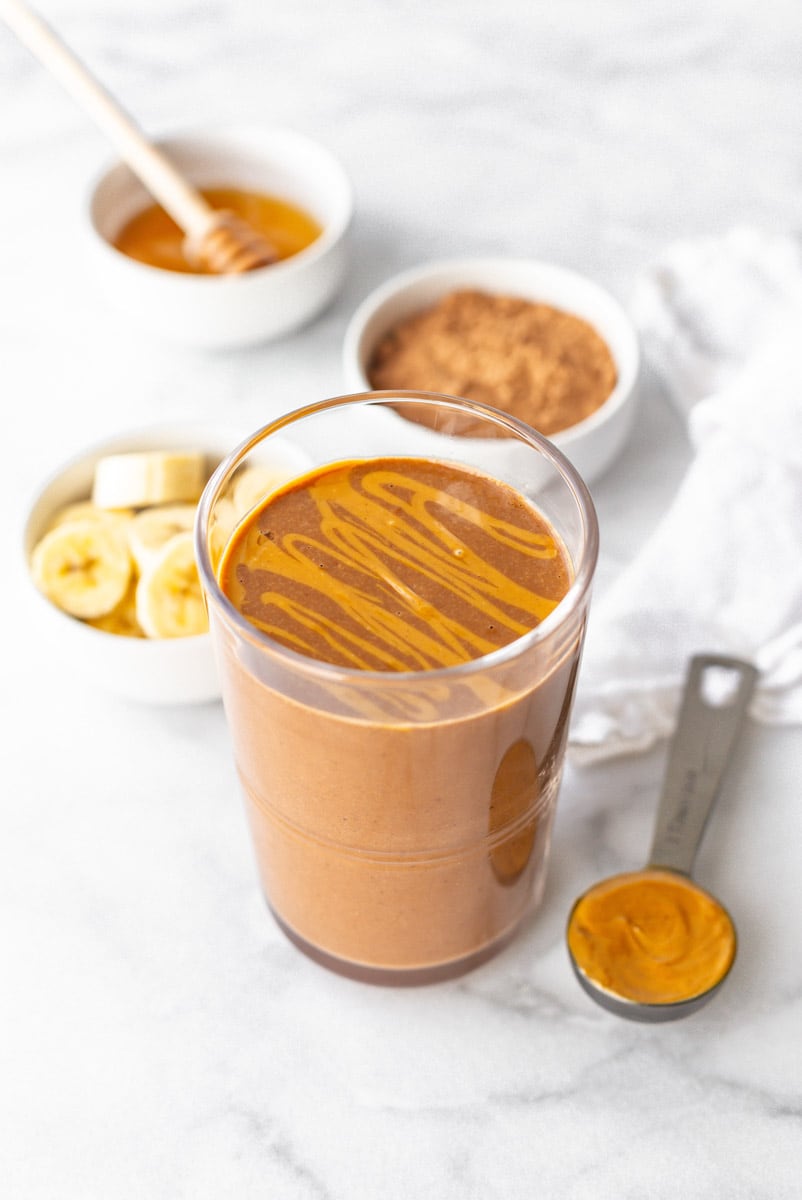 Finished smoothie in a glass with a peanut butter drizzle on top sit in front of small dishes filled with smoothie ingredients.