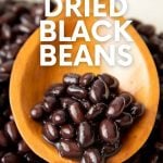 Seasoned black beans rest on a wooden spoon in a large pot of cooked beans. A text overlay reads, "How to Cook Dried Black Beans."