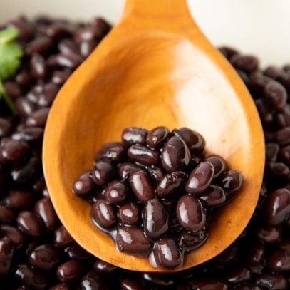 Seasoned black beans rest on a wooden spoon in a large pot of cooked beans.