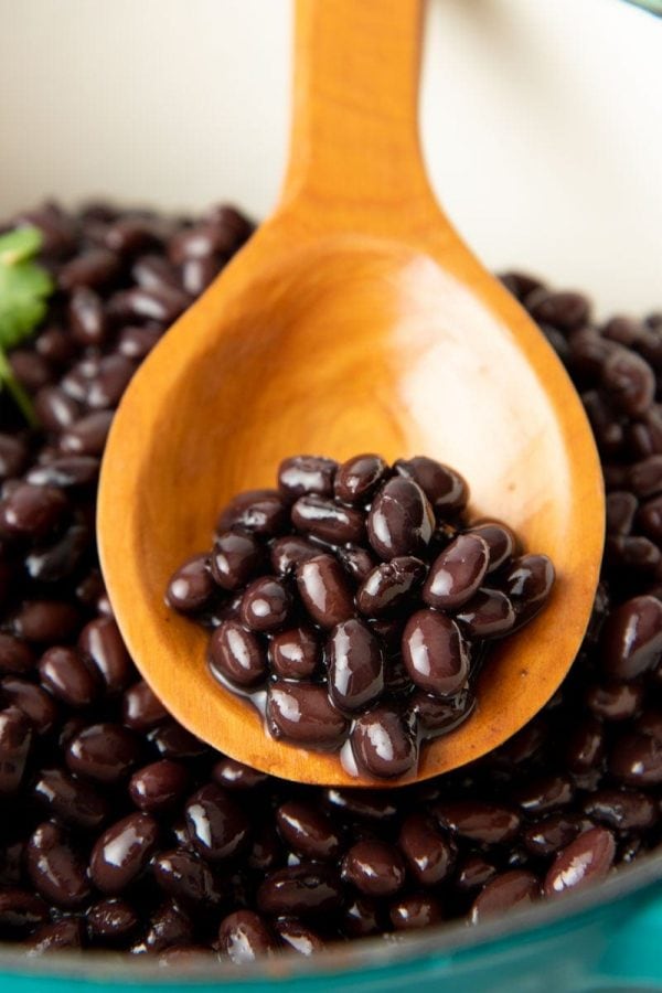 Seasoned black beans rest on a wooden spoon in a large pot of cooked beans.