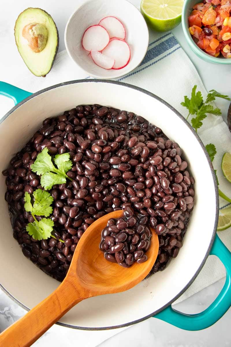 A wooden spoon scoops finished black beans out of a pot.