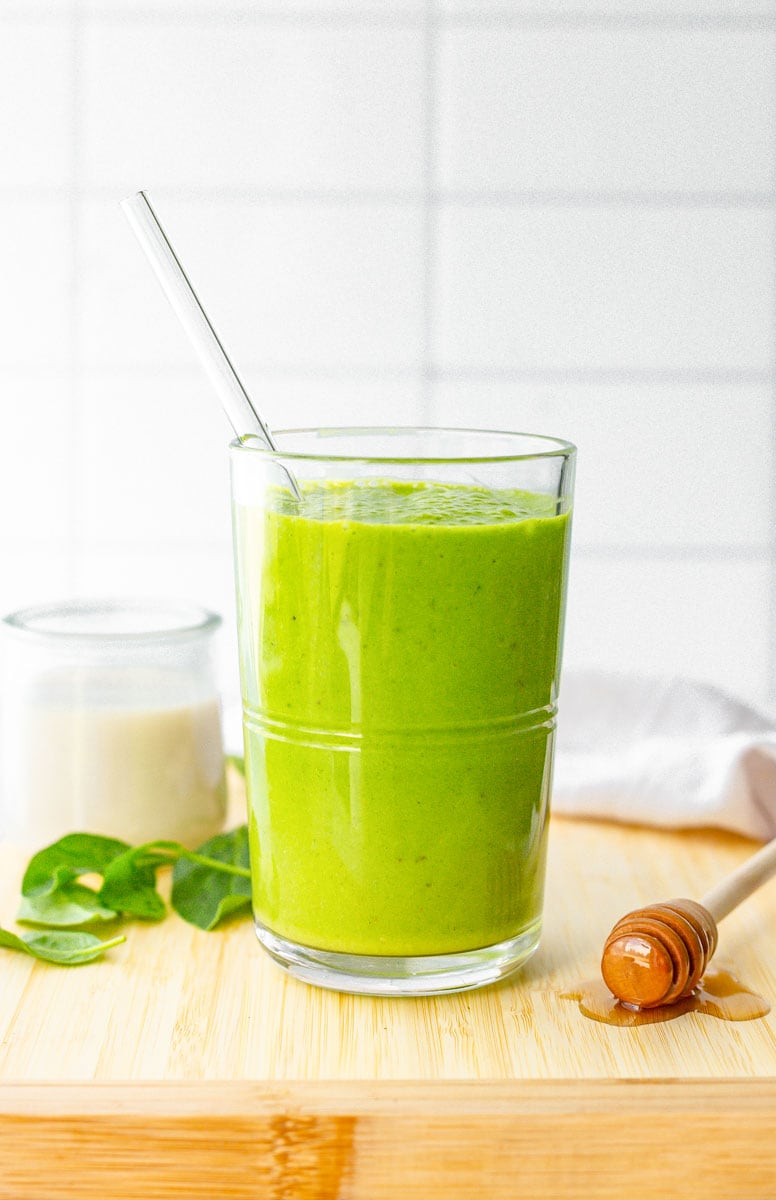 A glass filled with creamy green smoothie and a glass straw on a cutting board with fresh ingredients.