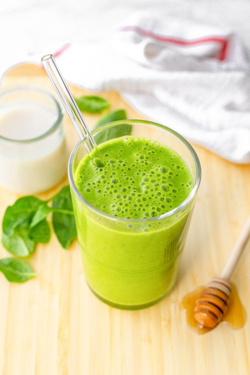 A green smoothie sits on a cutting boards with a honey drizzler and fresh spinach leaves beside it.