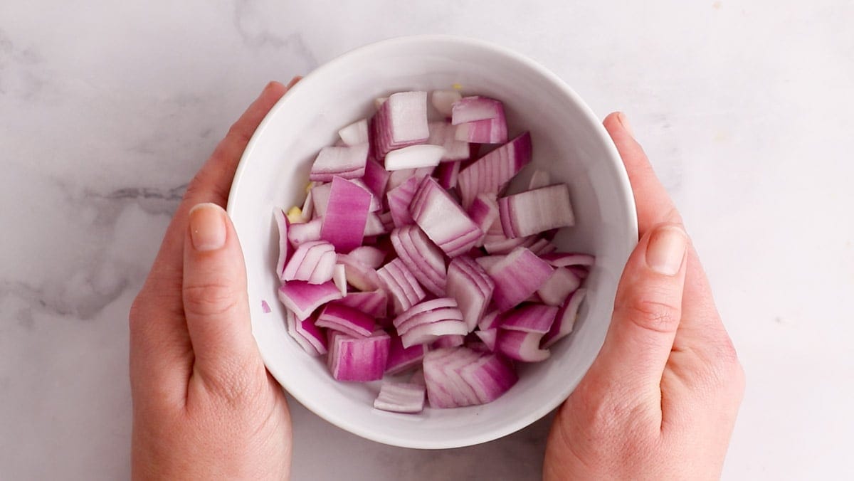 Overhead of hands holding a large white ramekin of chopped red onion.