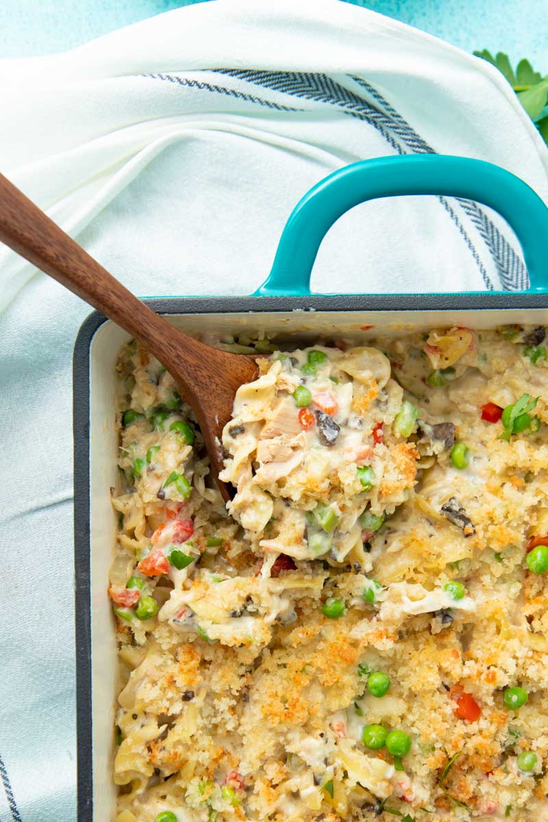 A wooden spoon scooping tuna noodle casserole out of a baking dish.