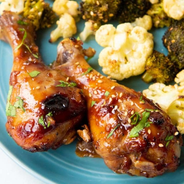 Close-up of two sticky chicken drumsticks on a plate with a side of roasted vegetables.