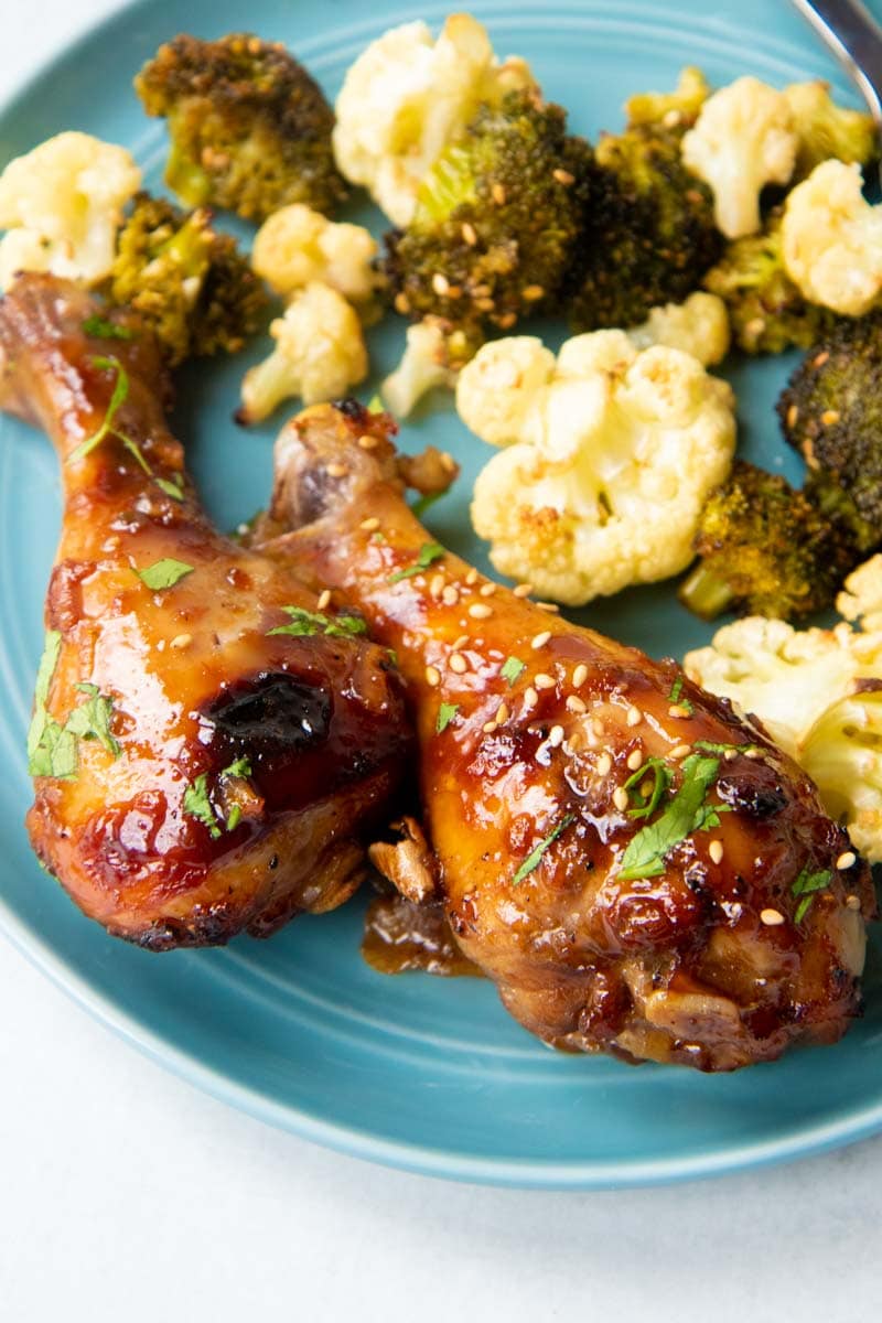 Close-up of two sticky chicken drumsticks on a plate with a side of roasted vegetables.