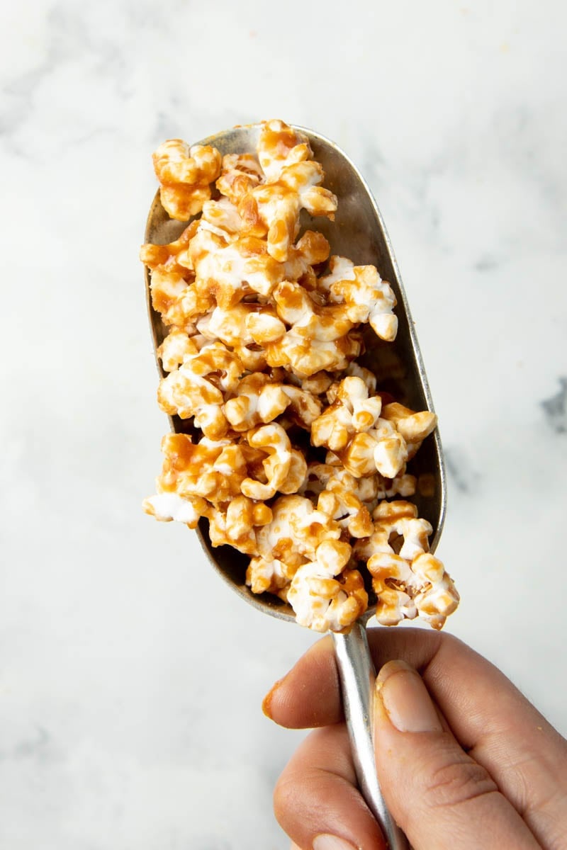 Overhead of a hand holding a metal popcorn scoop filled with finished caramel corn.