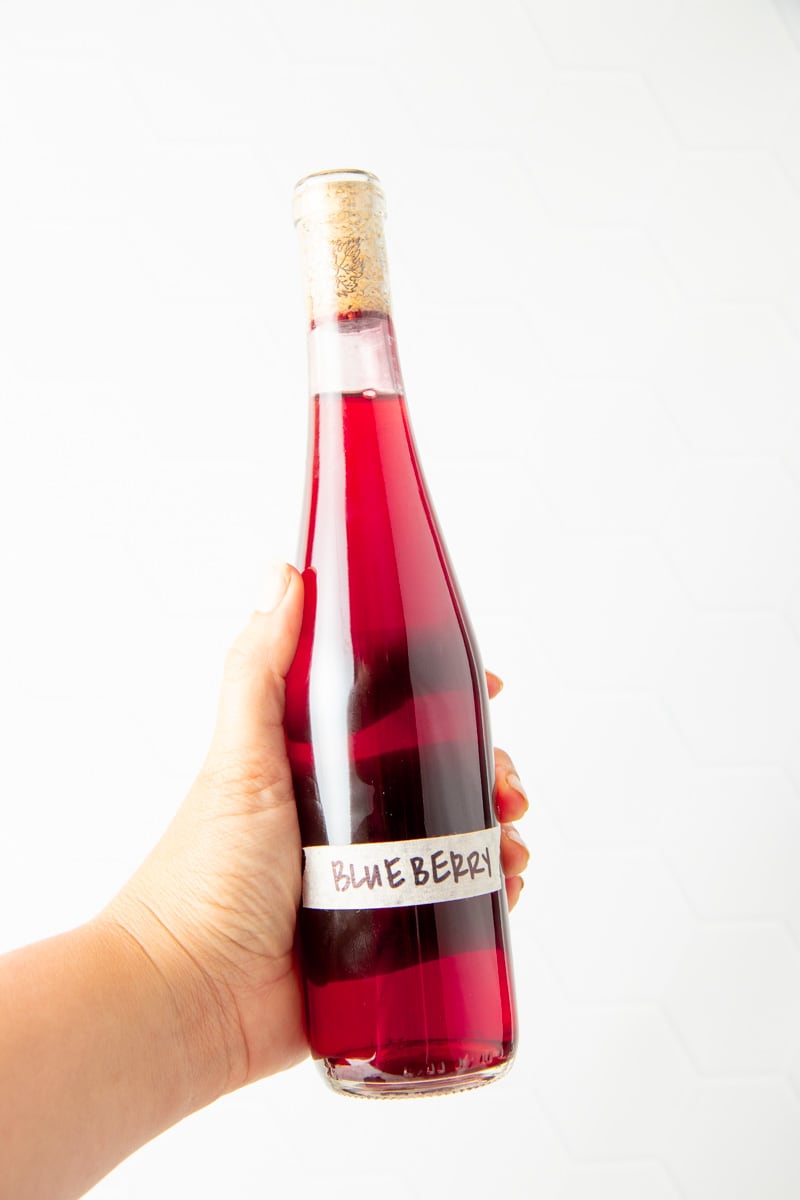 A hand holds a bottle of homemade blueberry wine sealed with a cork with a masking tape label that reads, "Blueberry."