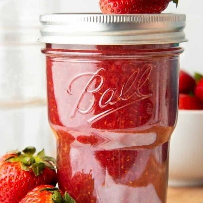 Strawberry jam in a closed and capped jar. A fresh strawberry sits on top of the lid, and two more berries are next to the jar.