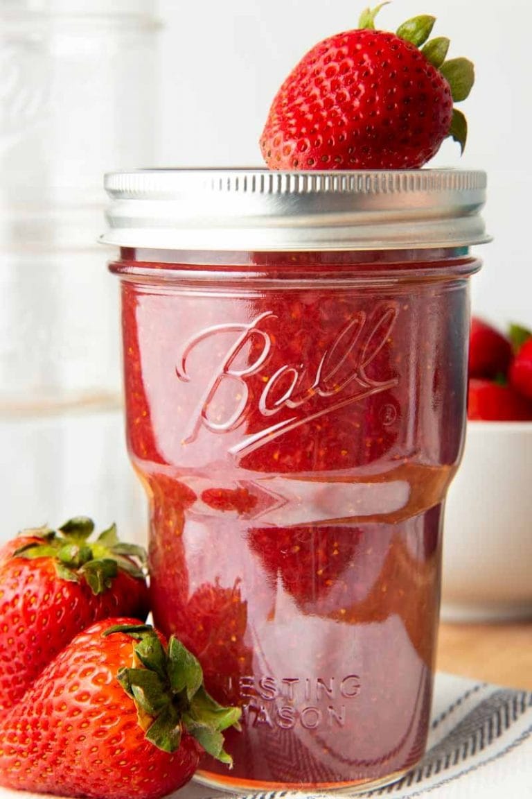Strawberry jam in a closed and capped jar. A fresh strawberry sits on top of the lid, and two more berries are next to the jar.