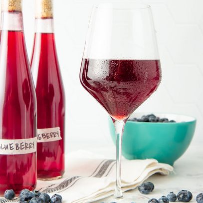 A stemmed wine glass filled with chilled blueberry wine stands surrounded by fresh blueberries and two bottles of homemade wine.