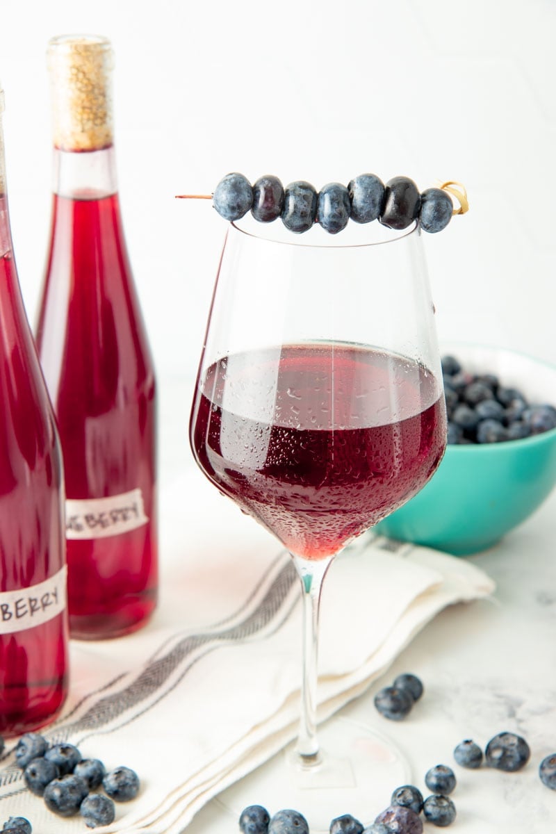 A cocktail skewer of fresh blueberries sits across the rim of a glass of homemade blueberry wine.