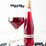 A chilled glass of blueberry wine with a cocktail skewer of fresh blueberries across the rim stands next to a bottle of homemade wine.