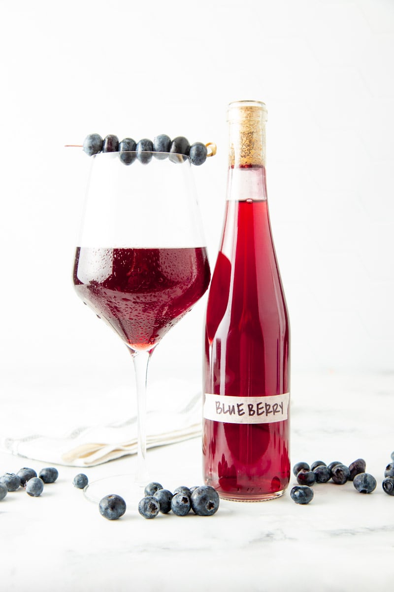 A chilled glass of blueberry wine with a cocktail skewer of fresh blueberries across the rim stands next to a bottle of homemade wine.