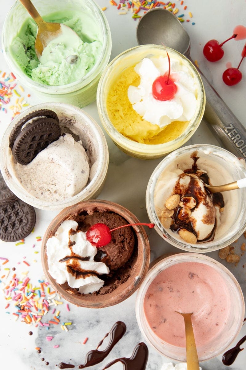 Six mason jars, each holding a different flavor of mason jar ice cream. An ice cream scoop, sprinkles, cherries, and chocolate cookies are on the table around the jars.