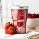 A full jar of strawberry jam sits on a folded tea towel in front of a bowl of strawberris.