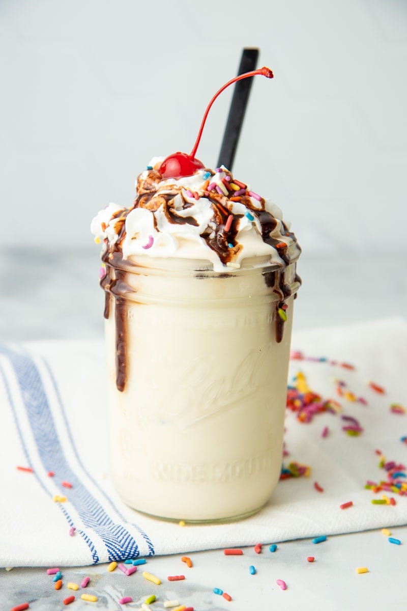 A jar of vanilla mason jar ice cream is topped with whipped cream, sprinkles, chocolate sauce, and a cherry. A spoon sticks out from the ice cream.