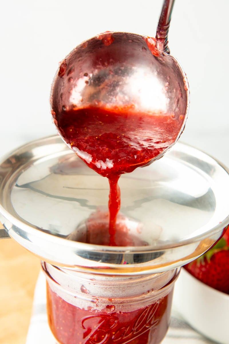 A metal ladle pours strawberry jam through a metal funnel into a glass jar